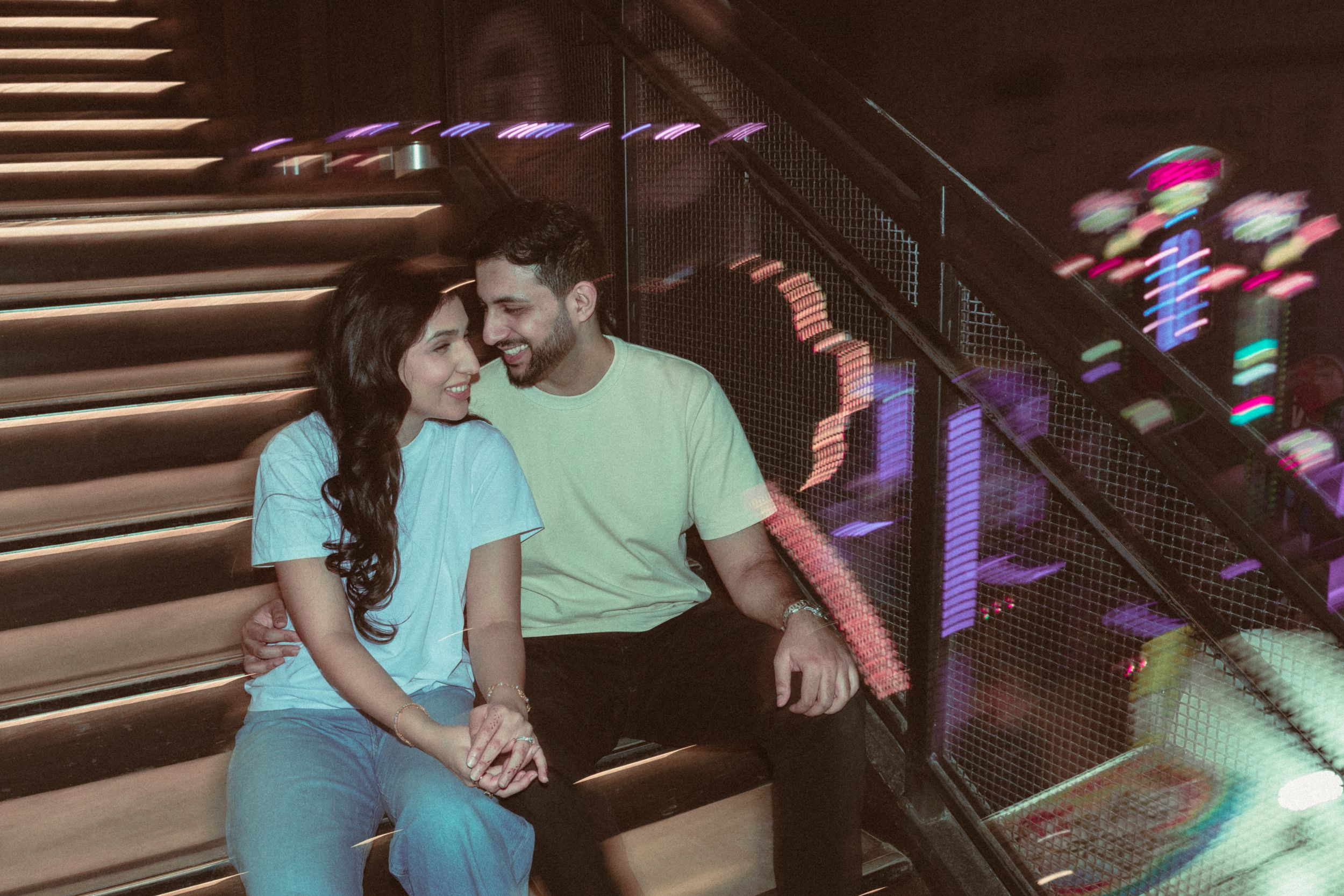Romantic couple sitting on stairs holding hands, smiling at each other in a colorful neon-lit venue.