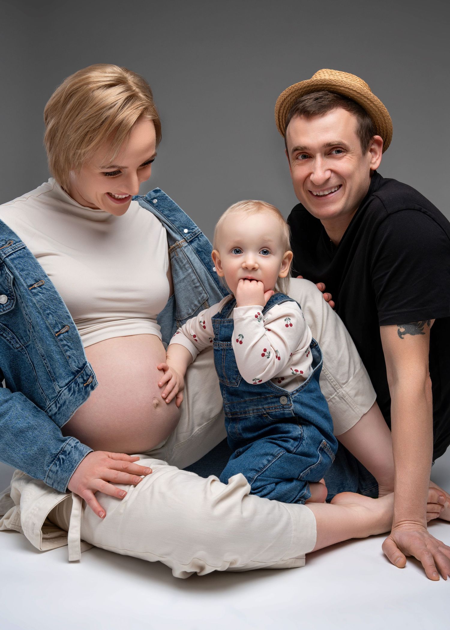 a family of three sits for a maternity photo shoot with the toddler resting a hand on their moms baby bump