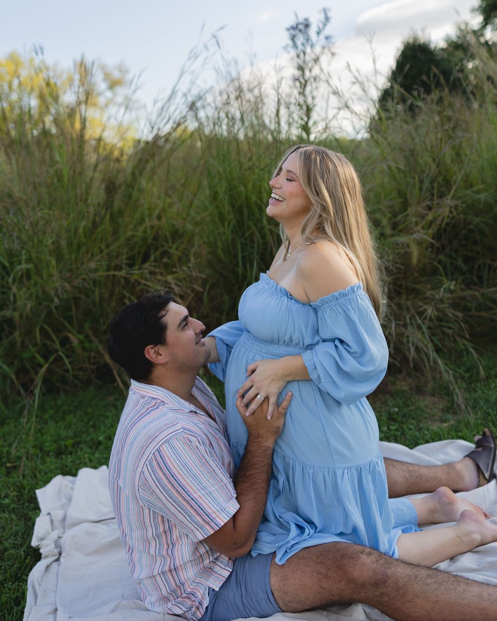 A couple sits together on a blanket outdoors, sharing a tender moment in a natural setting with tall grass and greenery.