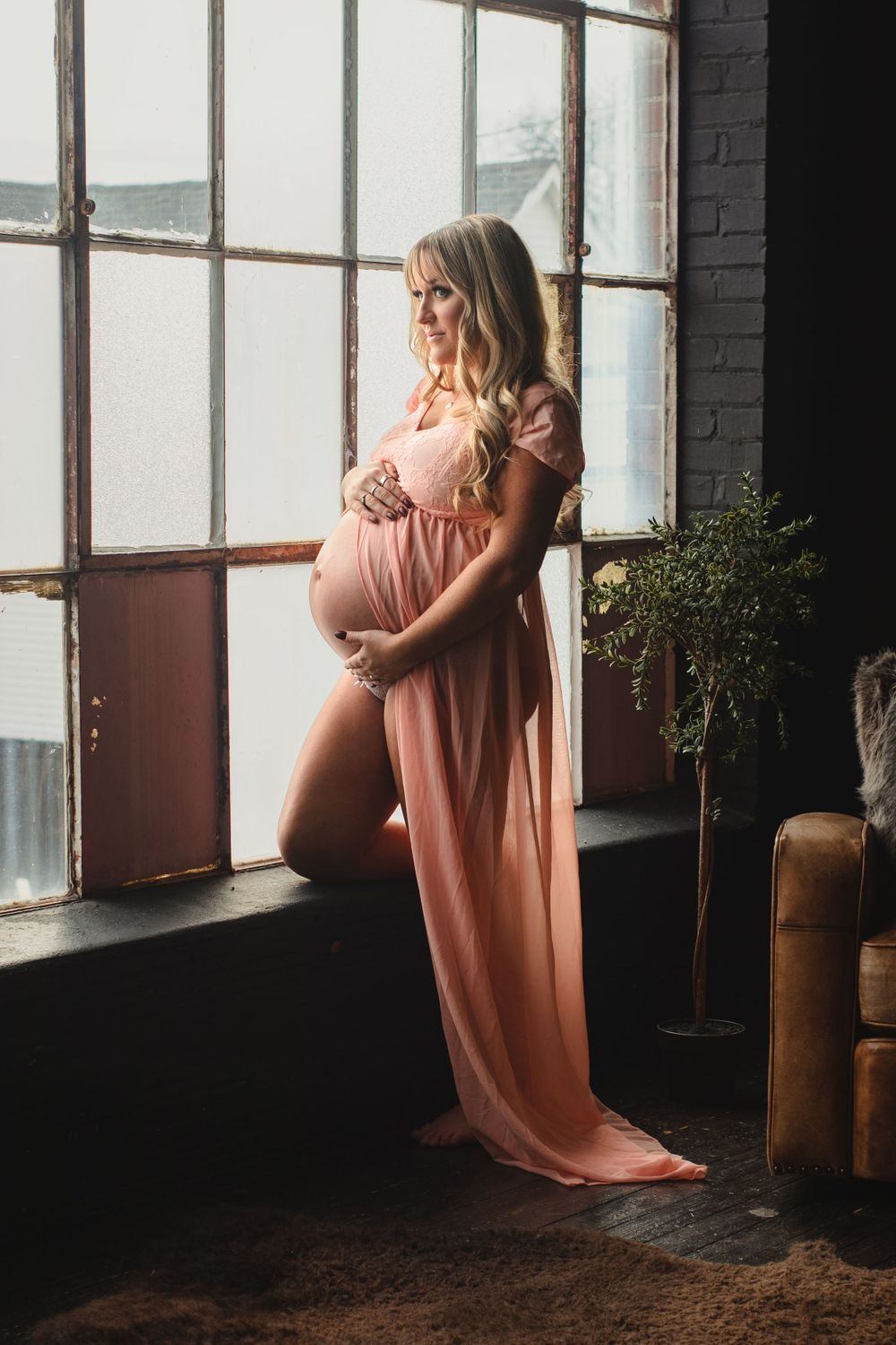 Expectant mother in flowing pink dress poses by industrial window.
