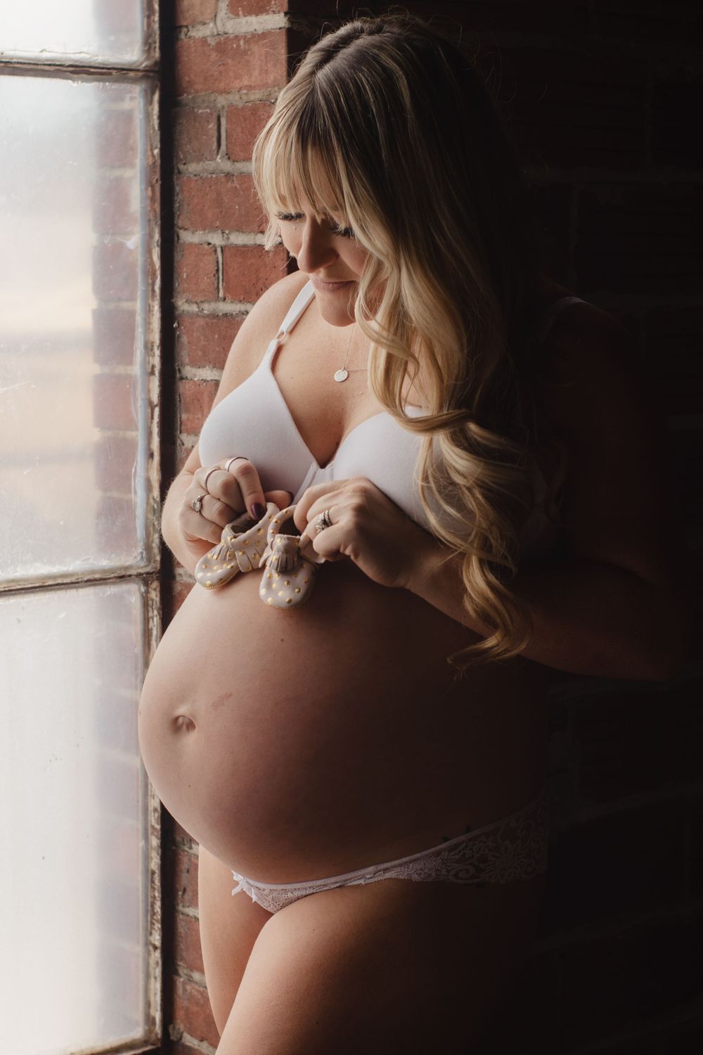 Maternity portrait by brick window showing woman in white lingerie.