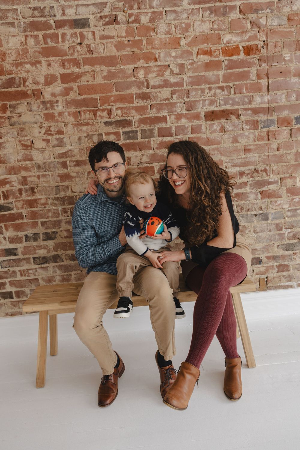 Happy family of three posing on a wooden bench against an exposed brick wall, parents smiling with toddler on lap.