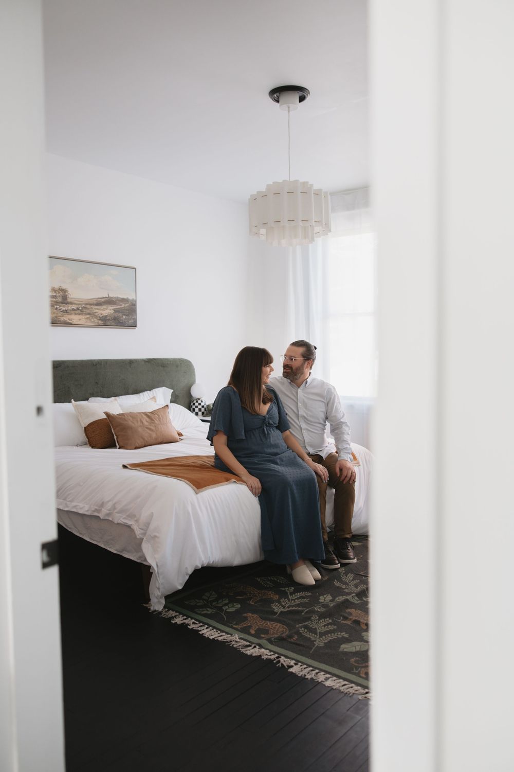 A couple sits together on a white bed in a bright minimalist bedroom with dark flooring.