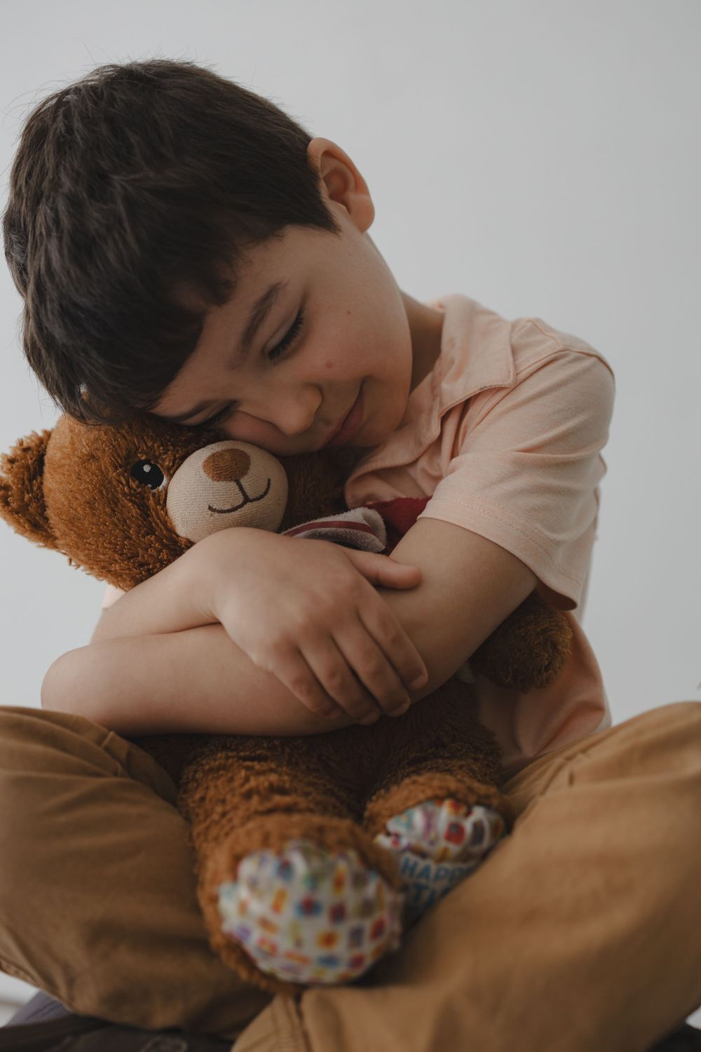 Young boy smiling and hugging a brown teddy bear with colorful patterned feet while sitting down.