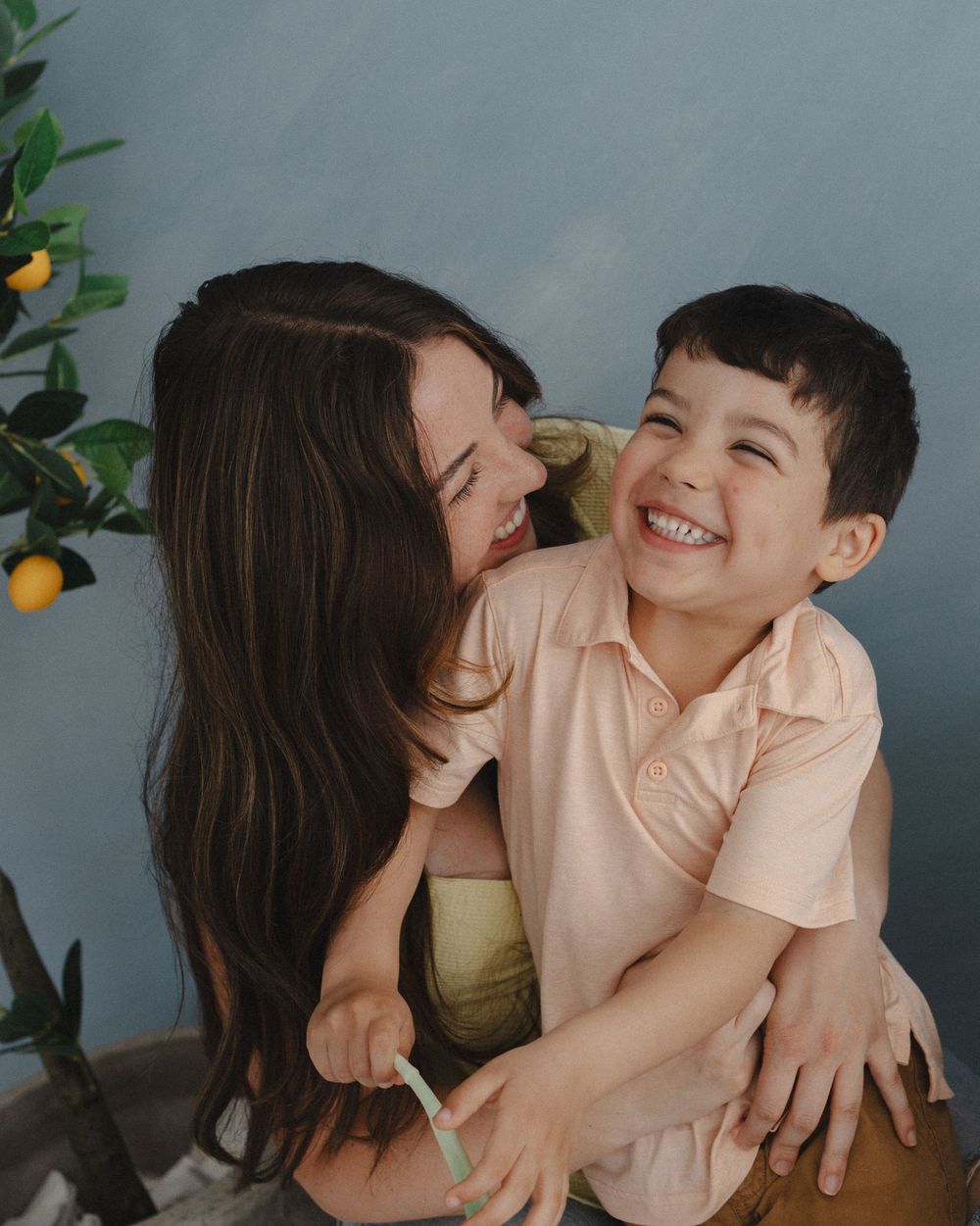 A joyful mother and young boy laughing together near a lemon tree against a blue background.