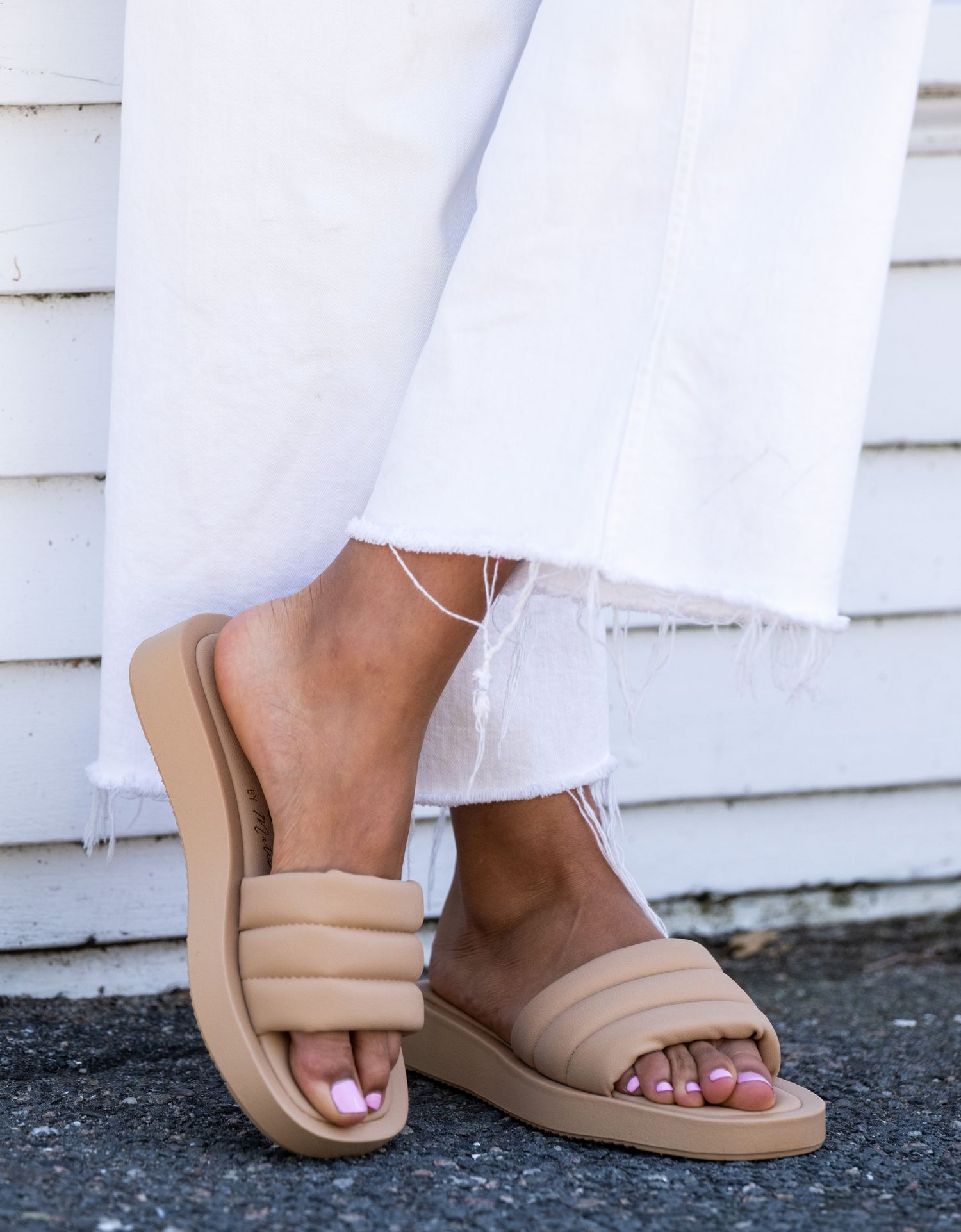 Close-up of feet wearing nude sandals against white dress and dark ground.