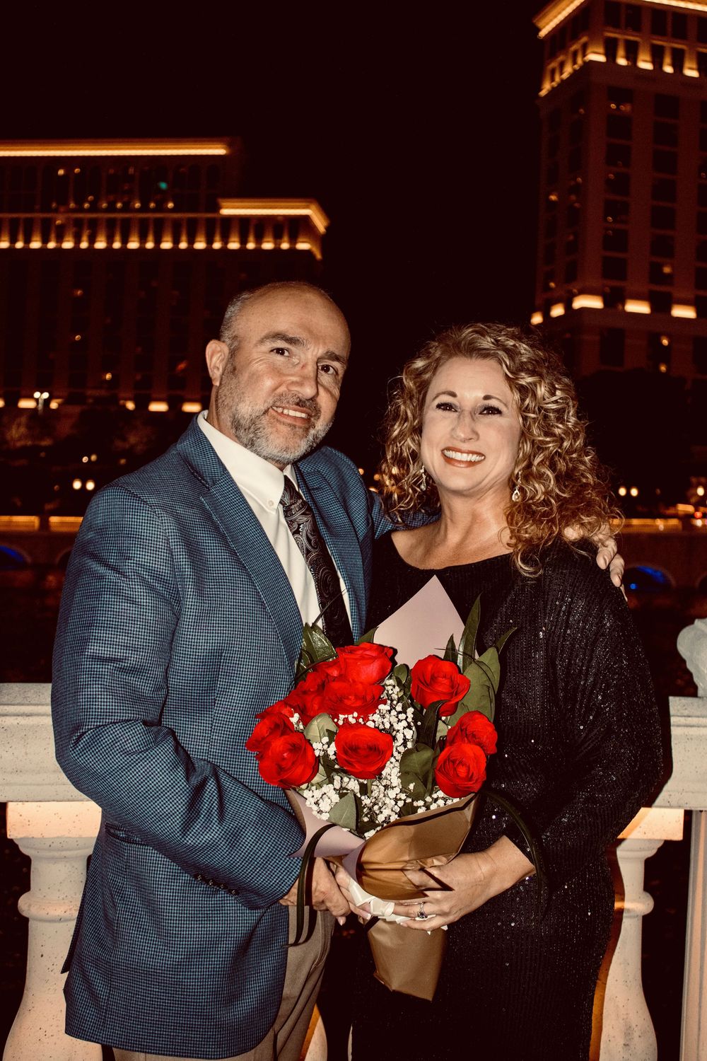 Smiling couple holding red roses bouquet at night with Caesars Palace lit up in the background.