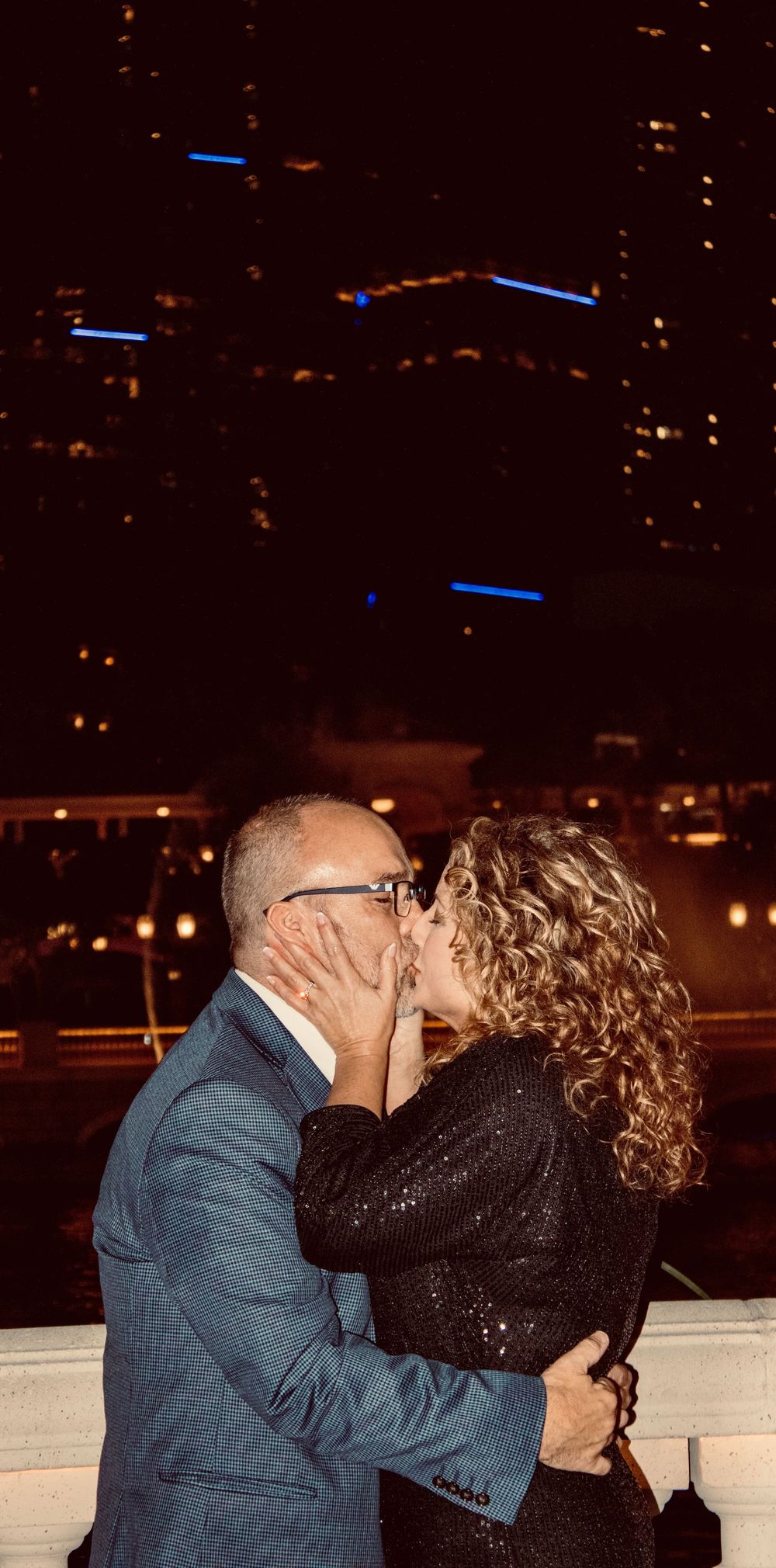 Newly engaged couple shares a kiss on a balcony with Las Vegas city lights glowing behind them.