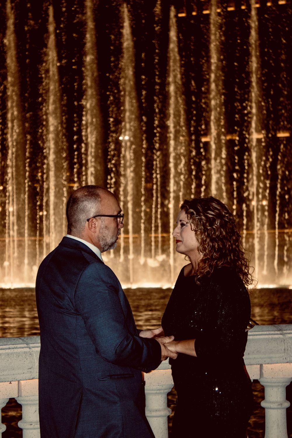 Couple holding hands facing each other in front of the Bellagio fountains at night.