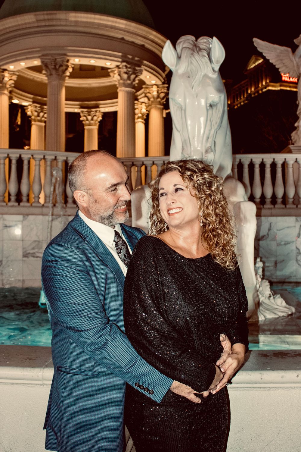 Romantic couple gazing at each other beside an illuminated fountain statue at Caesars Palace at night.