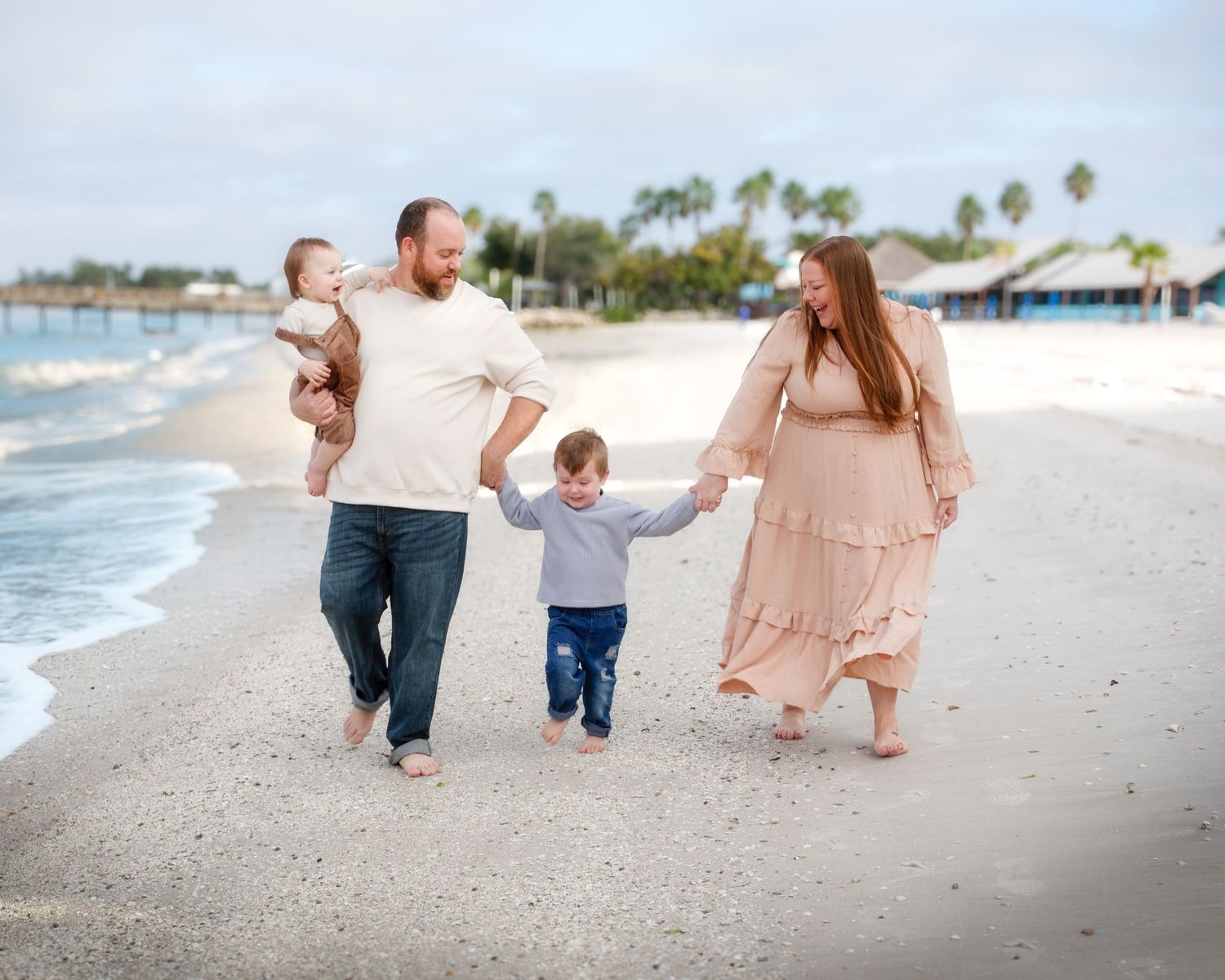 A family walks hand in hand along a beach with palm trees and ocean waves in the background.