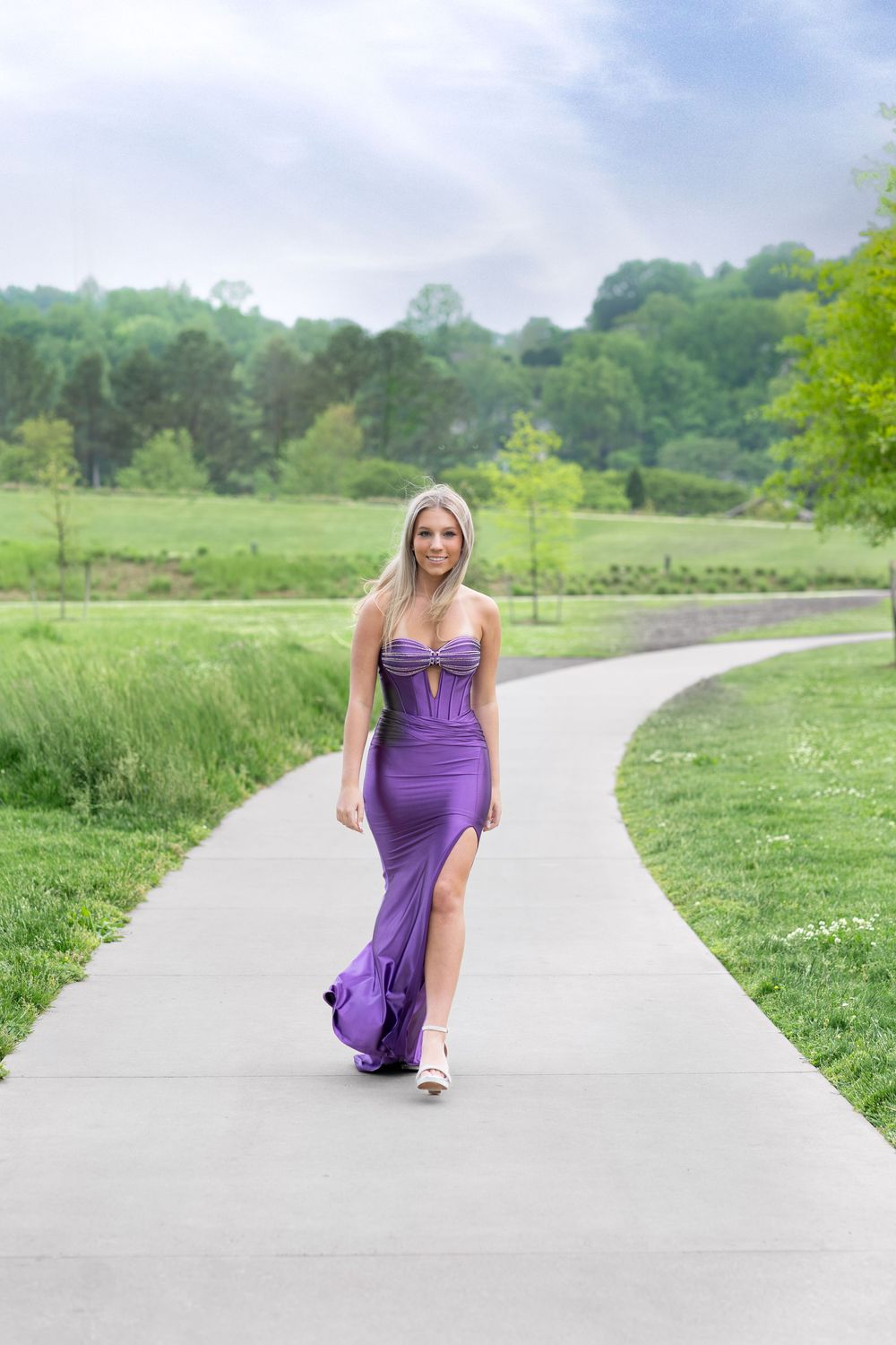 A person in a flowing purple gown walks along a winding concrete path surrounded by lush green grass and trees.