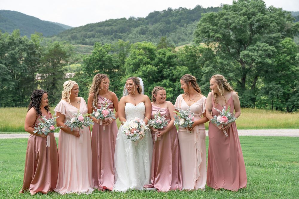 Wedding party in pink and mauve bridesmaid dresses pose together in a scenic outdoor field with mountains in background.