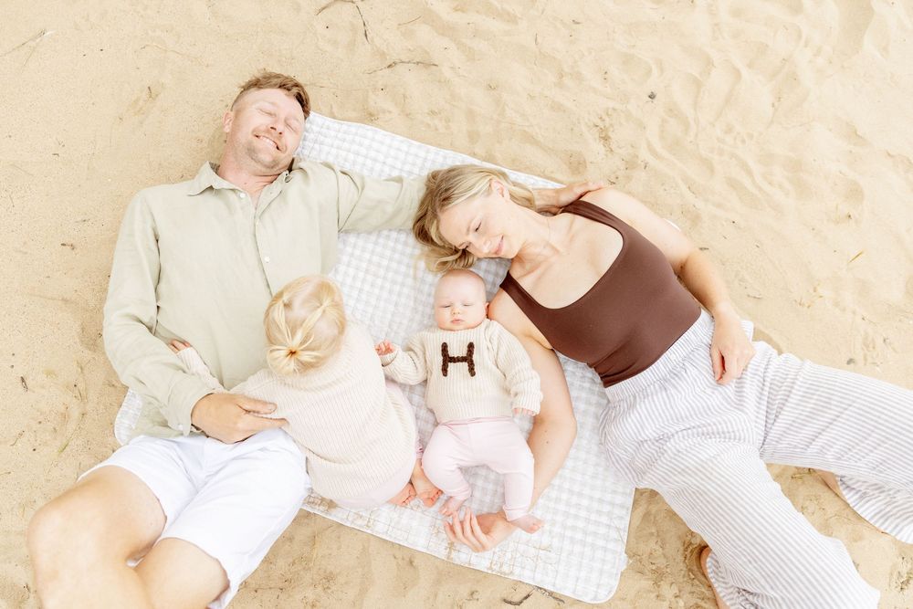 Family wearing white outfits lies together on beige blanket in natural outdoor setting during photo session.