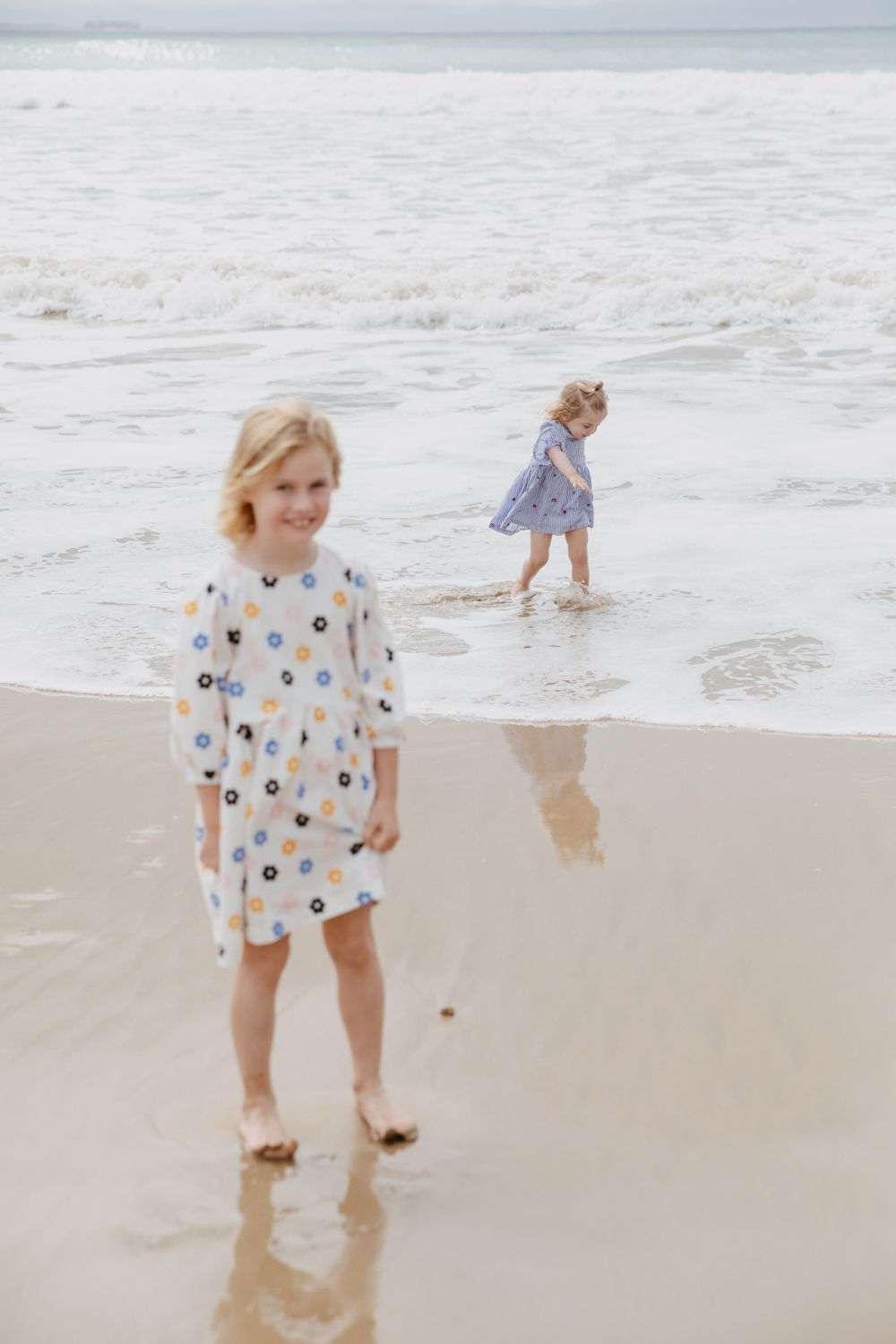 Two children in patterned dresses play at the edge of ocean waves on a sandy beach.