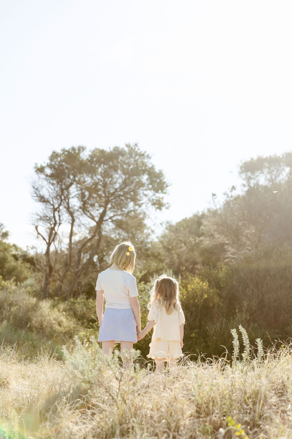 Two figures in light clothing hold hands while standing in a sunlit meadow surrounded by trees at golden hour.