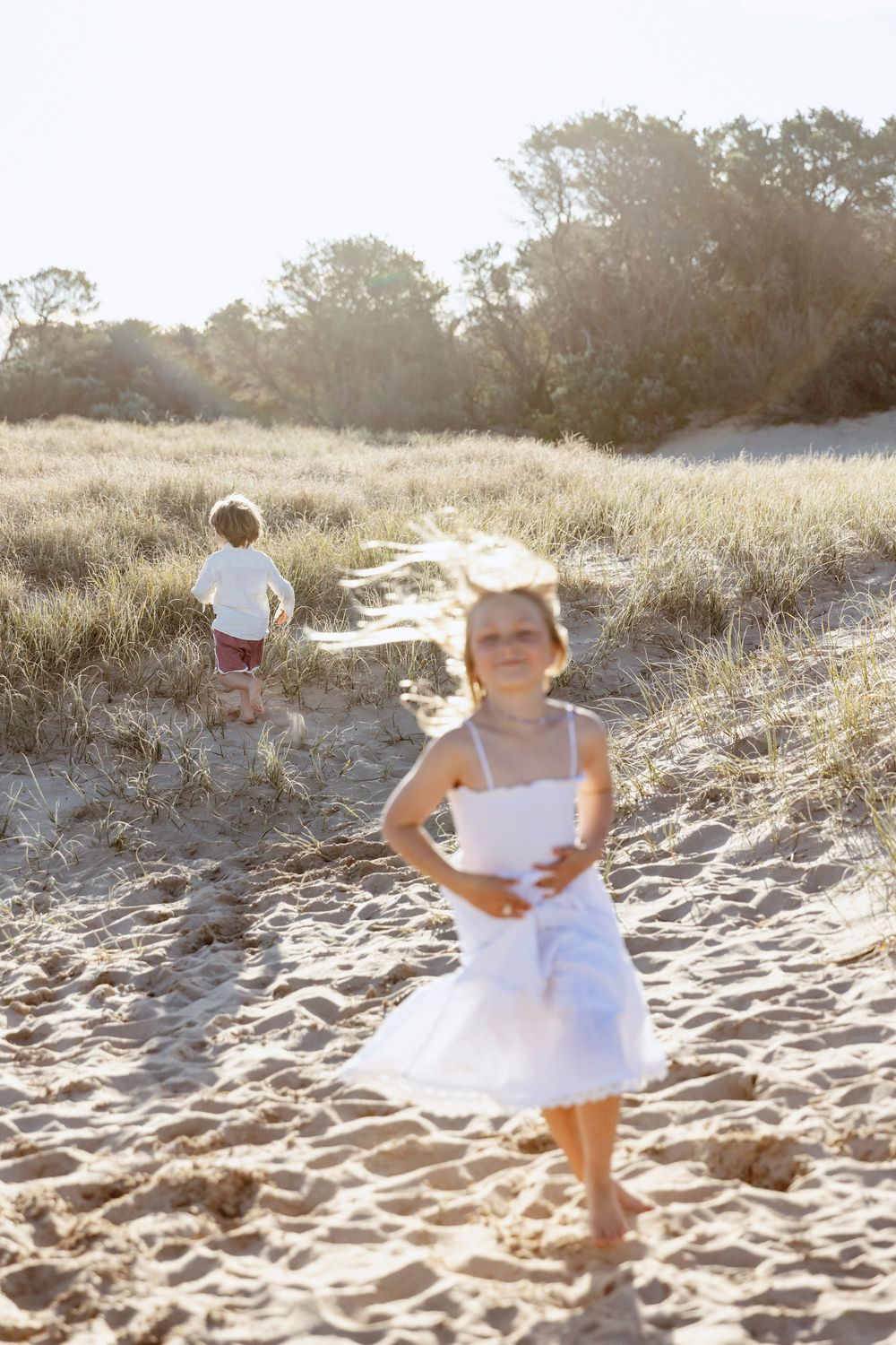 Child in white dress plays on sandy beach at sunset creating a dreamy summer scene.