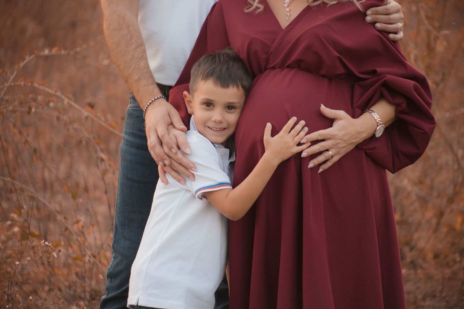 donna incinta con vestito scuro nel bosco , figlio maggiore la abbraccia, mani del bambino sul pancione della mamma. Sessione maternity