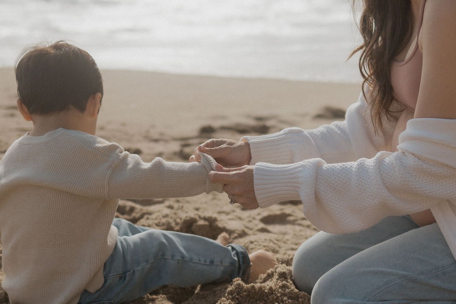 An adult rolls up the sleeve of a child's sweater on a sandy beach with the ocean blurred in the background.