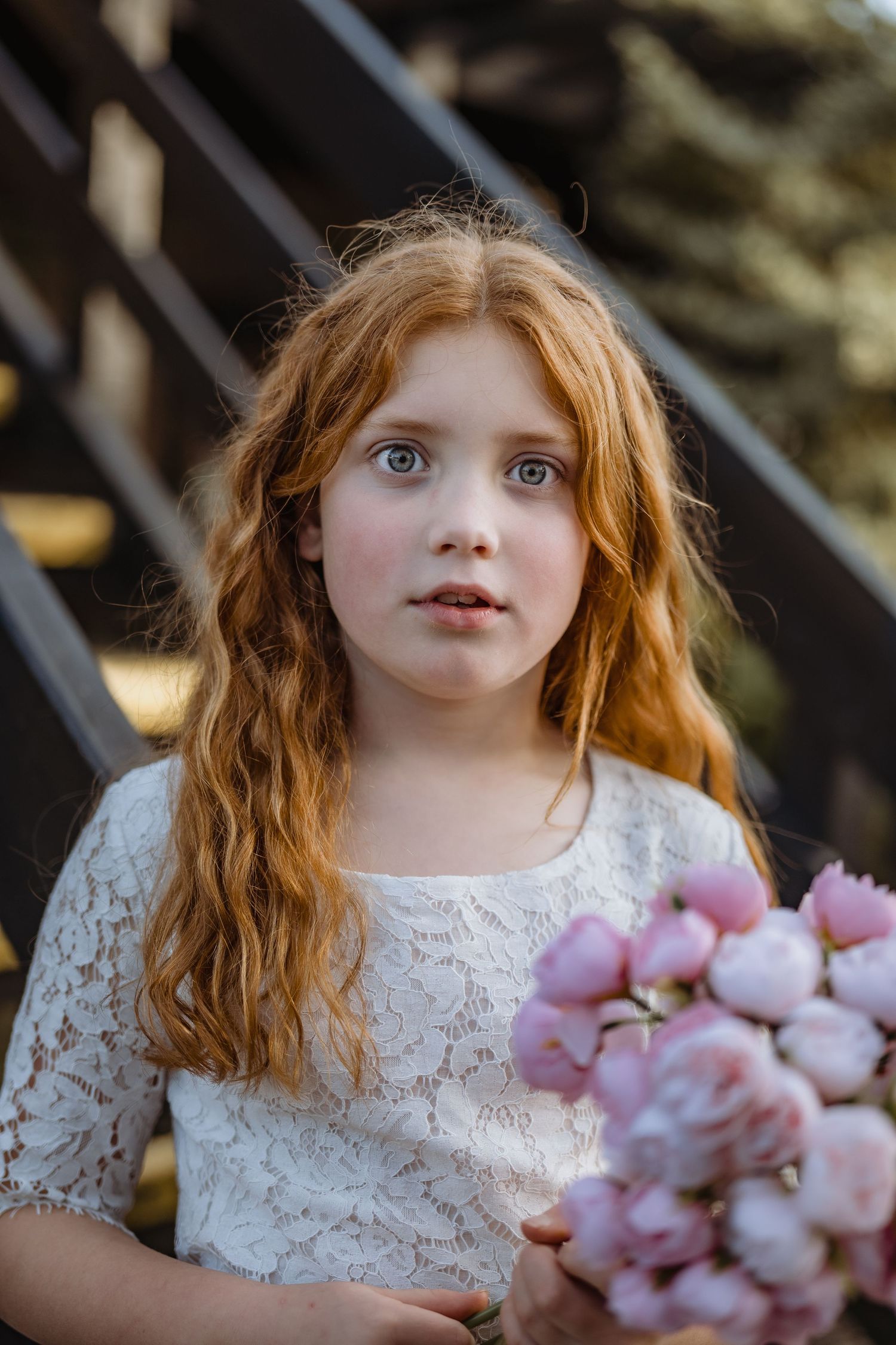 Young girl in white lace holding pink flowers outdoors.