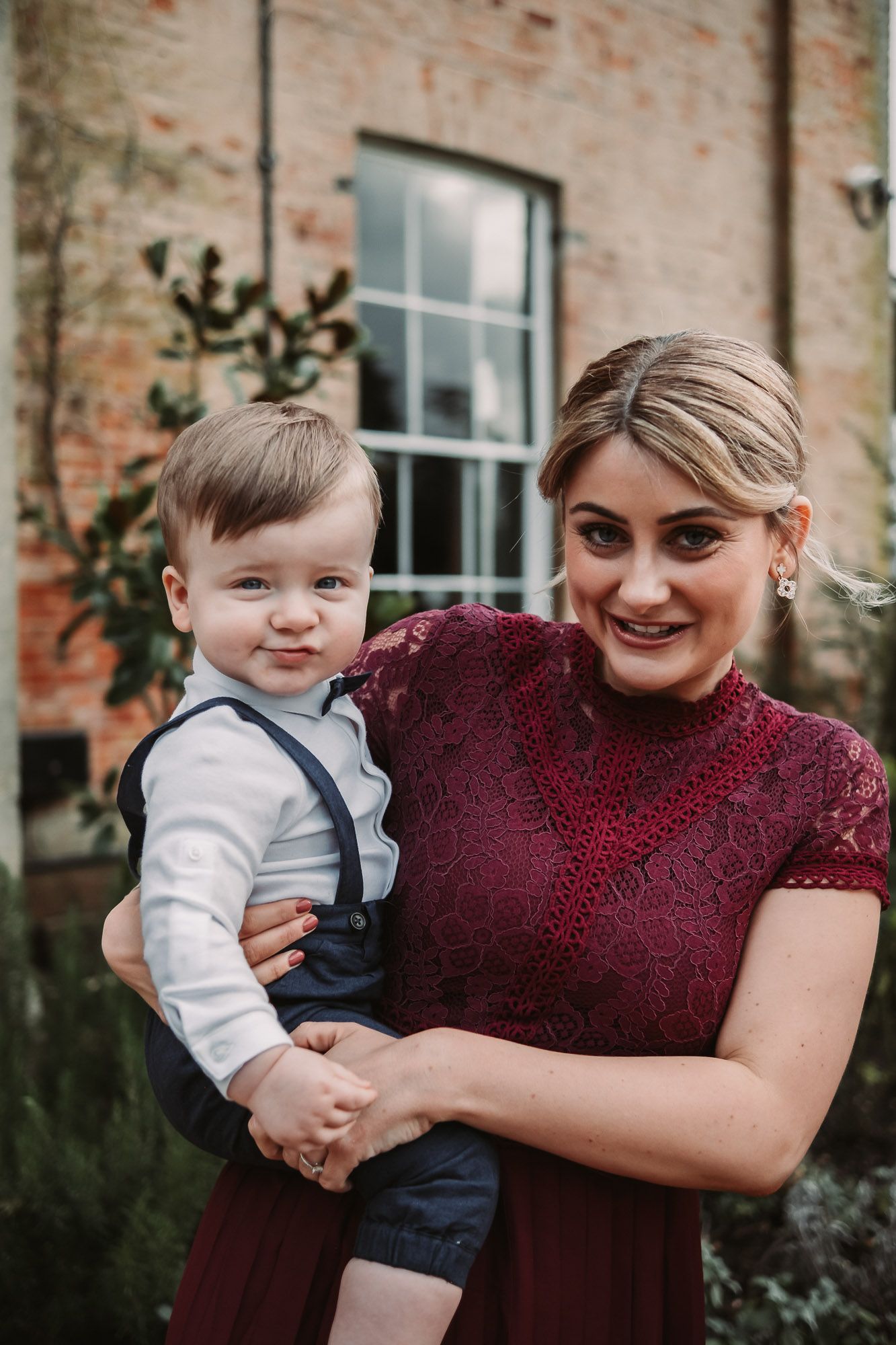 Woman in burgundy dress holding young child in formal attire at wedding.