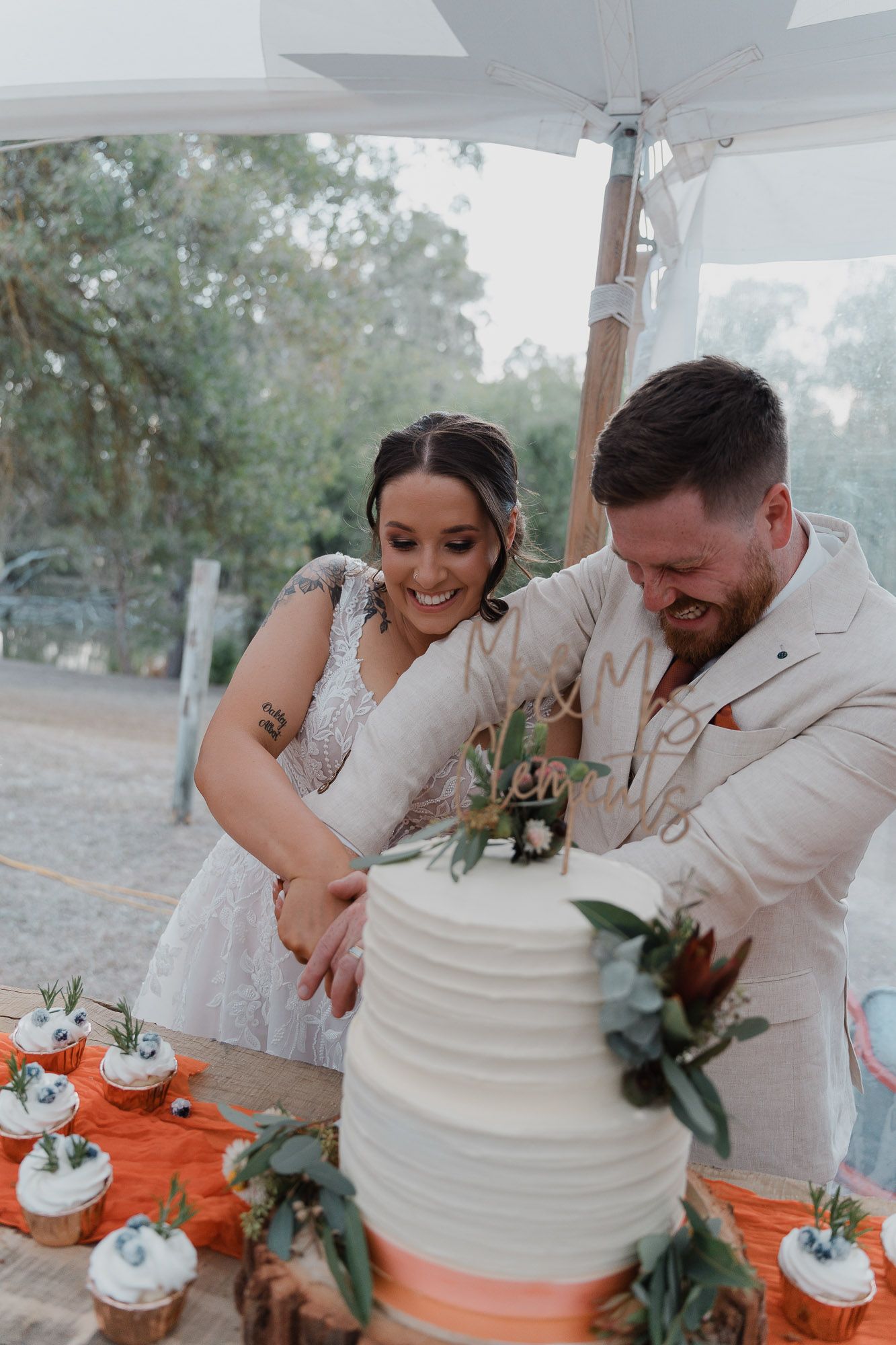 Couple cuts their white tiered wedding cake with greenery decorations.