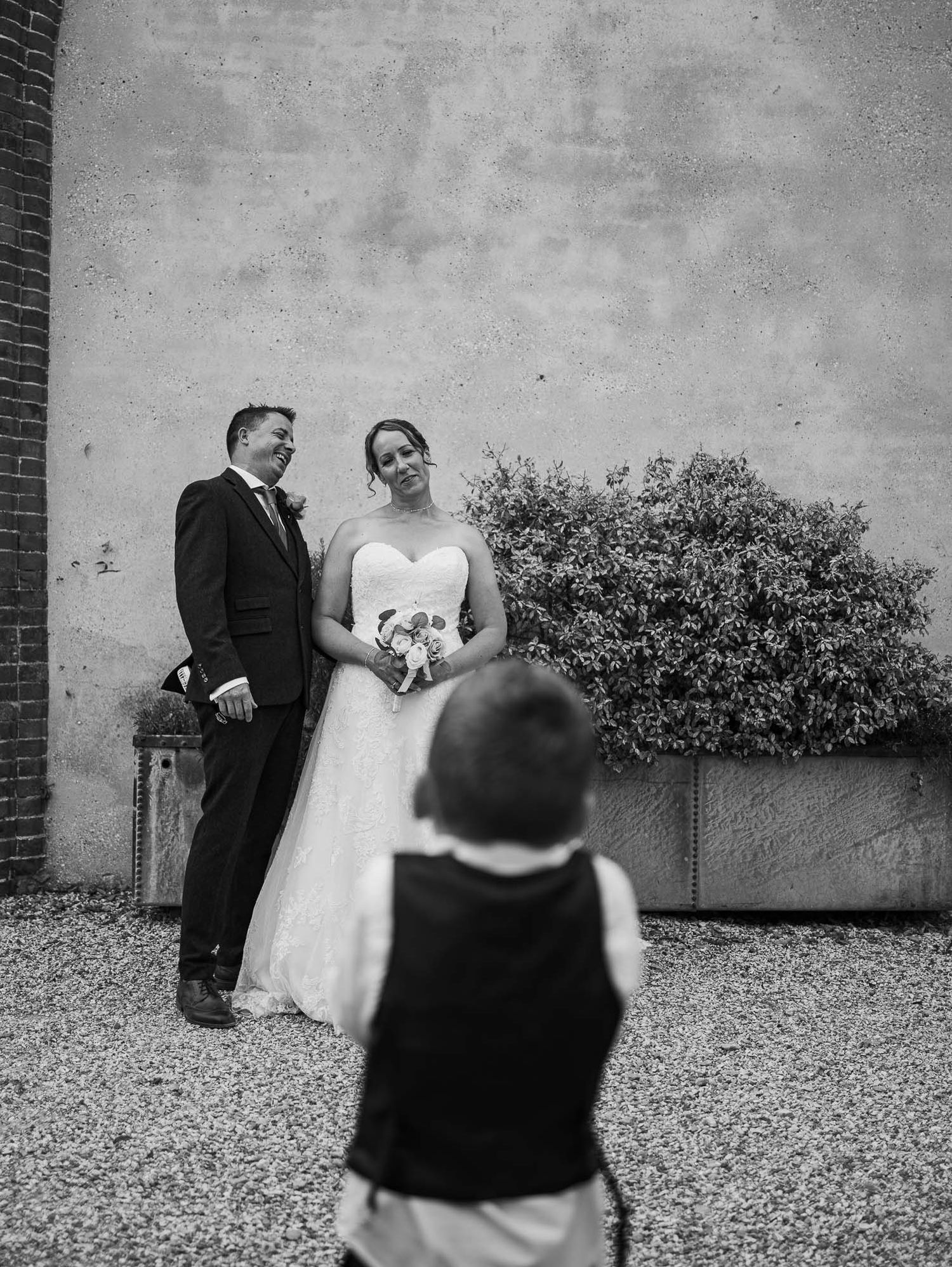 Black and white photo of a wedding couple posing against a brick wall with shrubs.