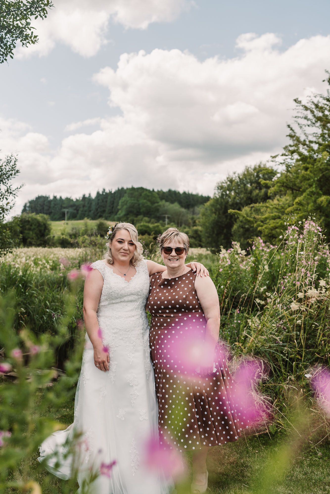 Two people posing in a sunny garden with purple wildflowers in the foreground.