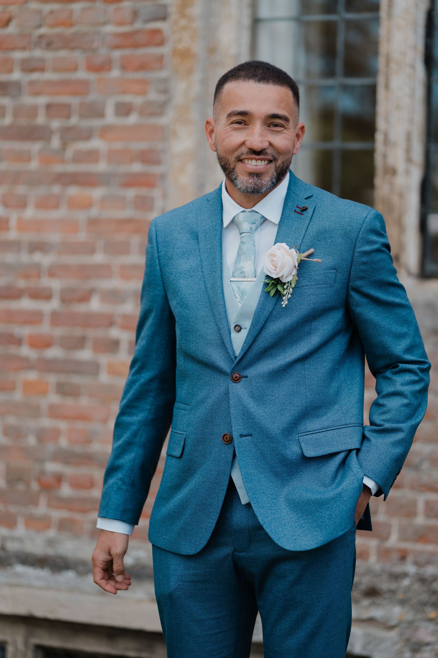 A groom in a stylish blue suit poses confidently against a brick wall.