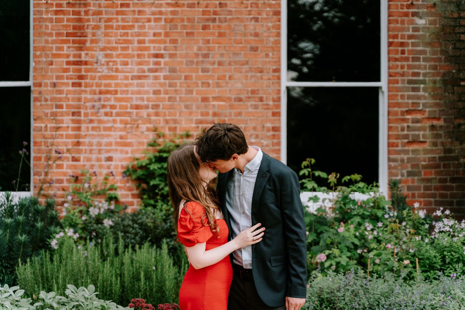 A couple poses by a brick wall with flowering garden herbs in the foreground.
