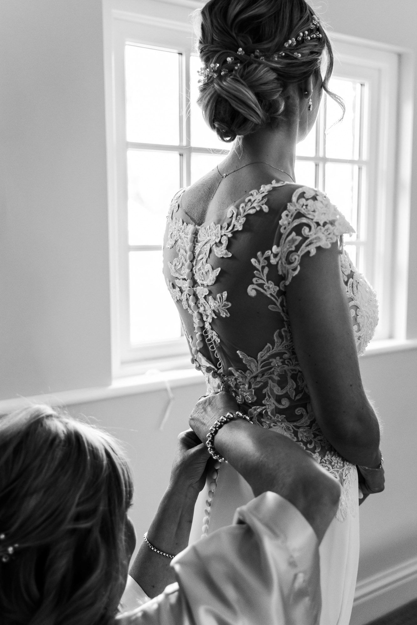 Elegant lace wedding dress being buttoned up from behind near a bright window.
