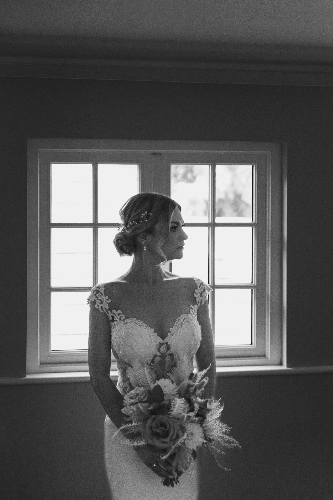 Silhouette of bride in lace dress standing by window holding bouquet.