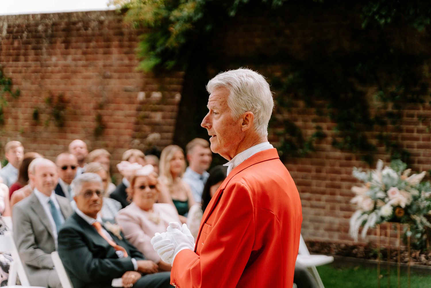 Speaker in bright orange jacket addresses outdoor wedding ceremony audience.