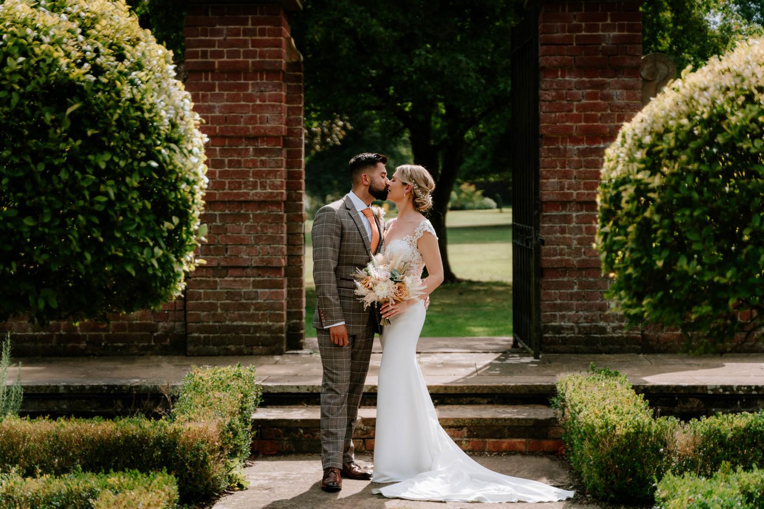Newlyweds share intimate moment between brick pillars in garden setting.