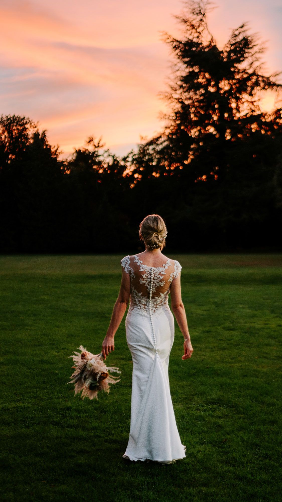 Bride in fitted lace gown holds bouquet during sunset in garden setting.