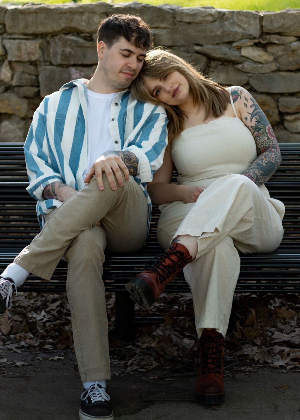 A couple in casual attire sits together on a bench near a stone wall.