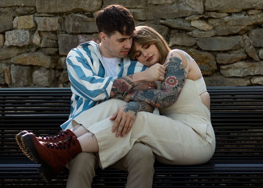 A couple shares a tender moment while sitting on a bench against stone architecture.