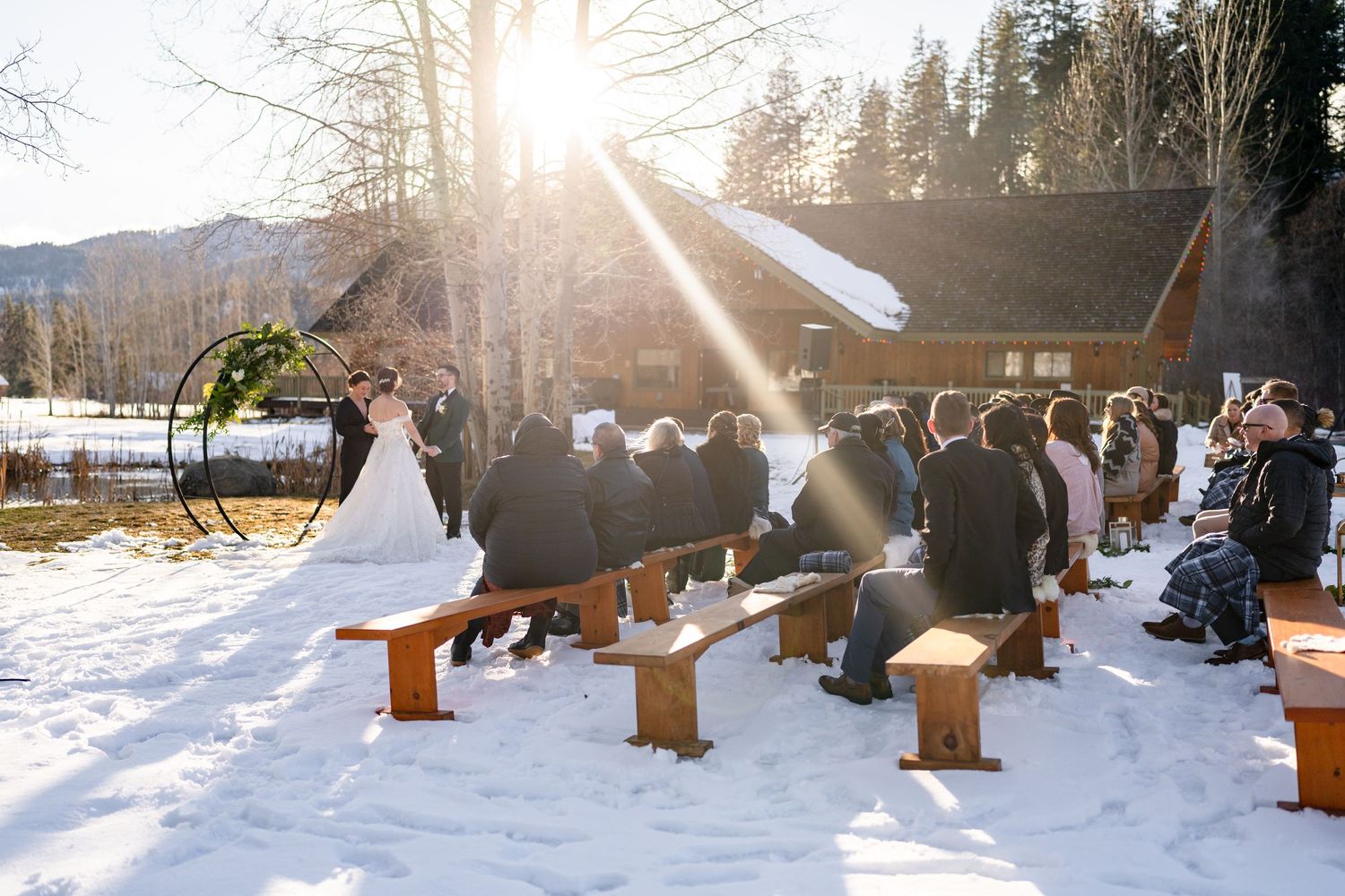 A winter outdoor wedding ceremony with guests seated on wooden benches in the snow as sunlight streams through trees.