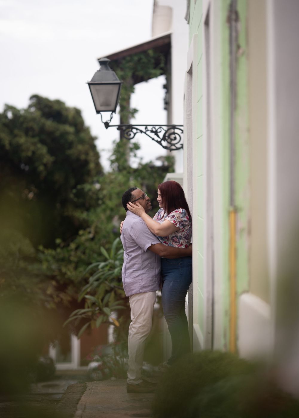 A romantic couple embraces under a vintage street lamp against a white building wall.