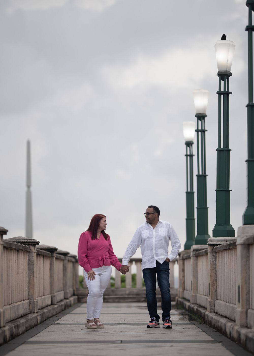 A couple holds hands while walking on a bridge with vintage lampposts.