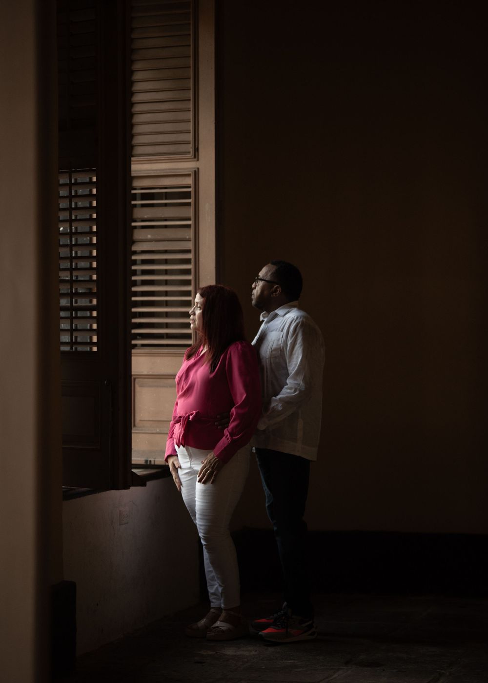 A couple stands by a window in dramatic lighting creating a silhouette effect.