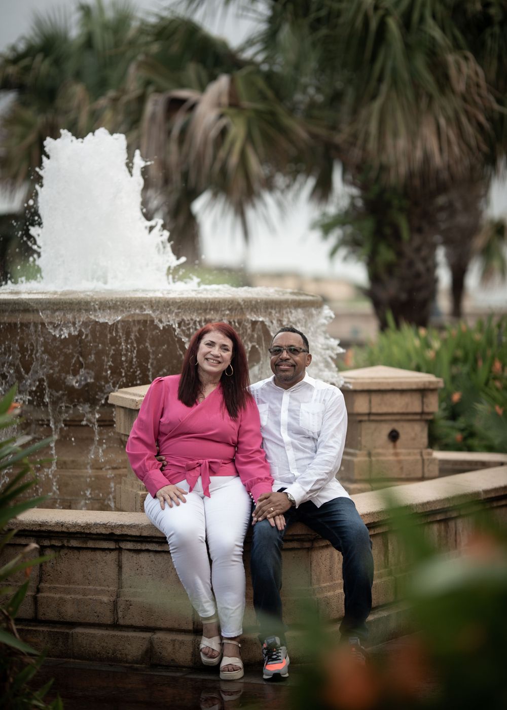 A couple sits together by a fountain with palm trees in the background.