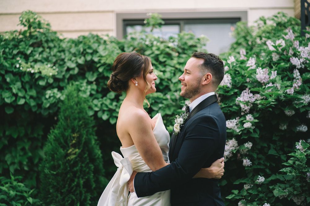 Newlyweds embrace in garden setting surrounded by blooming lilac bushes.