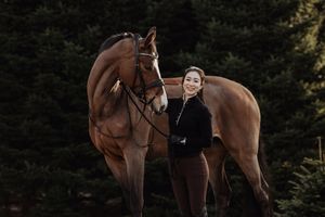 A smiling woman in black equestrian attire stands beside a chestnut horse in front of evergreen trees.