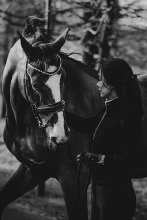 Black and white photo of a woman in equestrian gear bonding with a bridled horse in a forest setting.