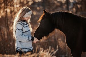 Young blonde girl in Nordic sweater gently feeds a dark horse in golden autumn light.
