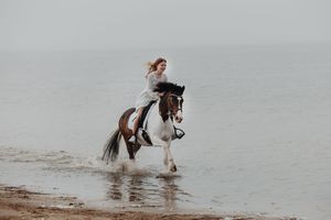 A woman rides a brown and white horse through shallow ocean waves on a misty beach.