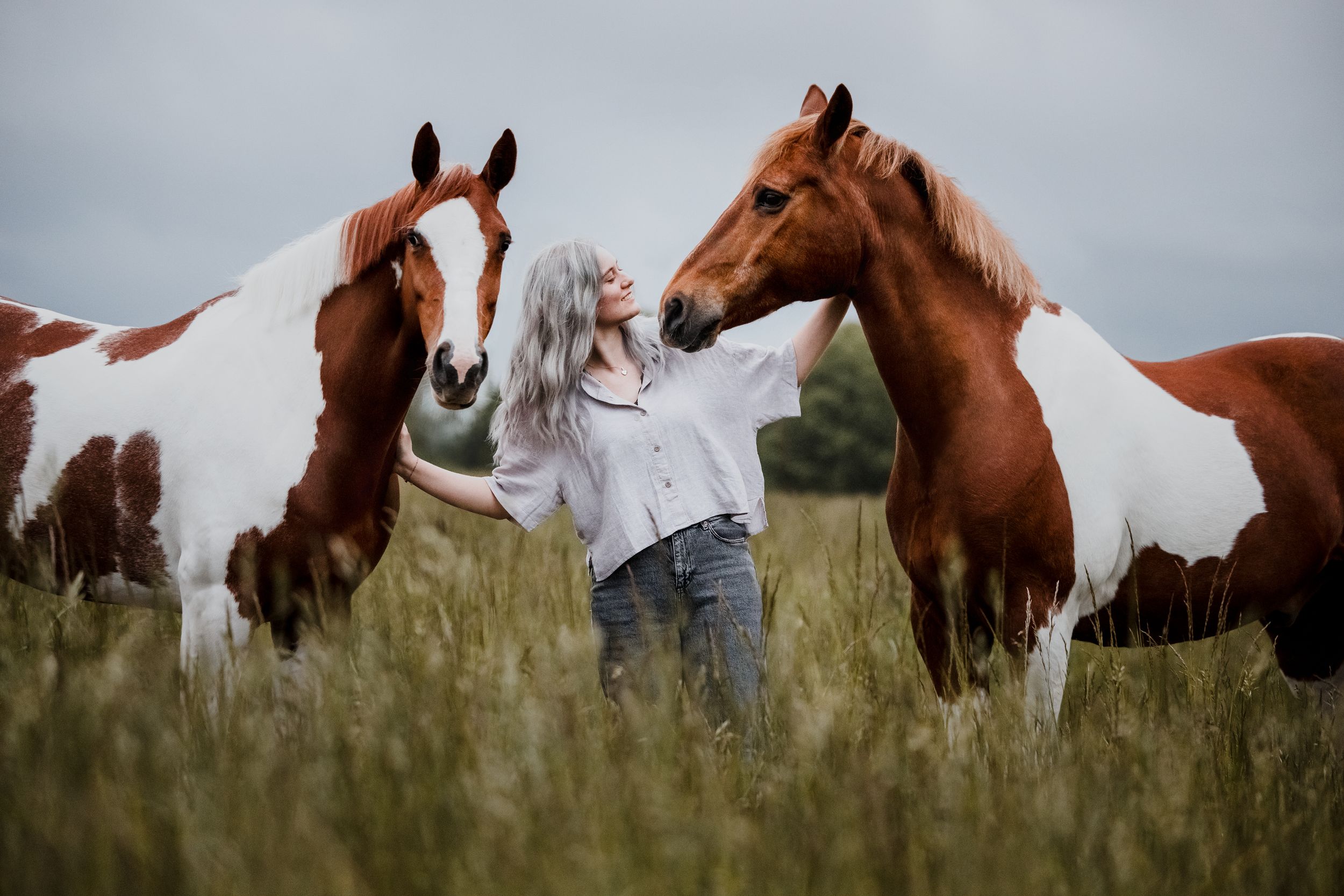 Woman with white hair stands between two brown and white paint horses in a tall grass field under cloudy skies.