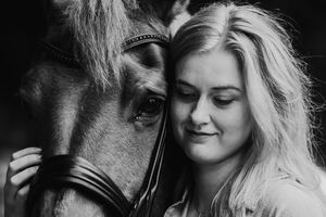 Black and white portrait of a young blonde woman tenderly resting her head against a horse wearing a bridle.