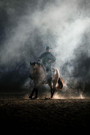 Cowboy riding a horse through dramatic smoke and dust in a dark arena, wearing a black hat and western gear.